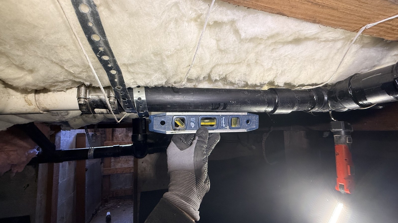 Gloved hand holding a blue level against a new black drain pipe under insulated floor joists, with a work light glowing in the crawlspace