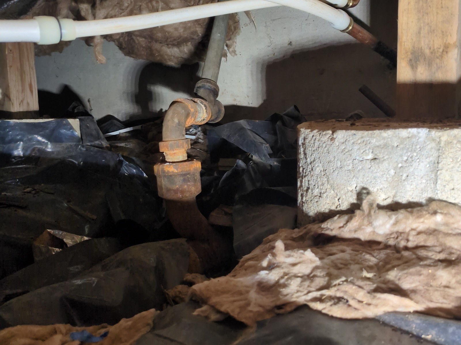 Corroded galvanized drain elbow and fittings in a dim crawlspace, white plastic water lines overhead and a concrete pier block at right
