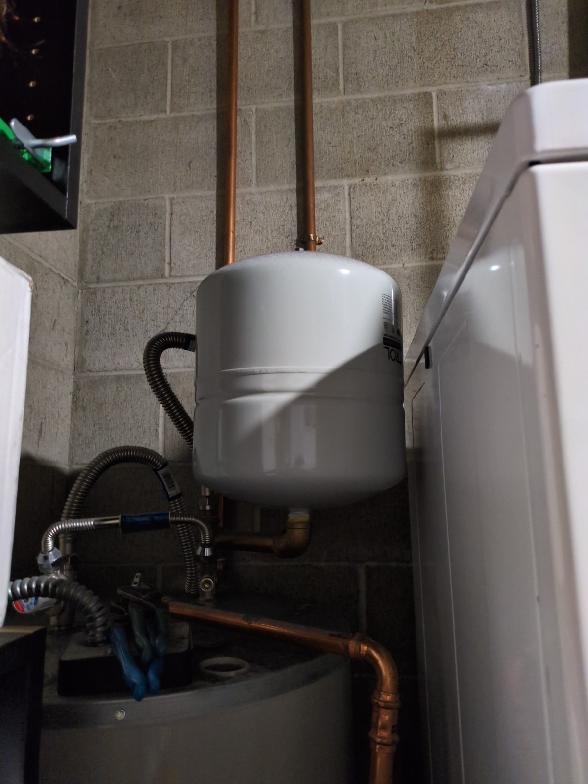 New white expansion tank mounted on copper piping above a water heater beside a laundry area