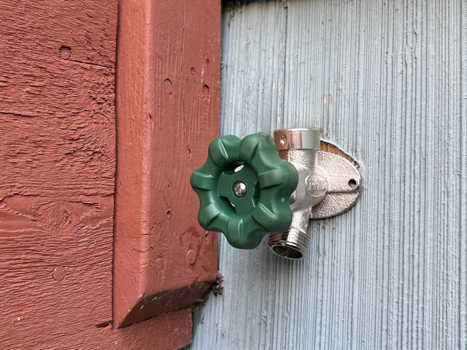 Chrome outdoor faucet with a green round handle mounted on blue-gray shingle siding next to a red-painted deck post