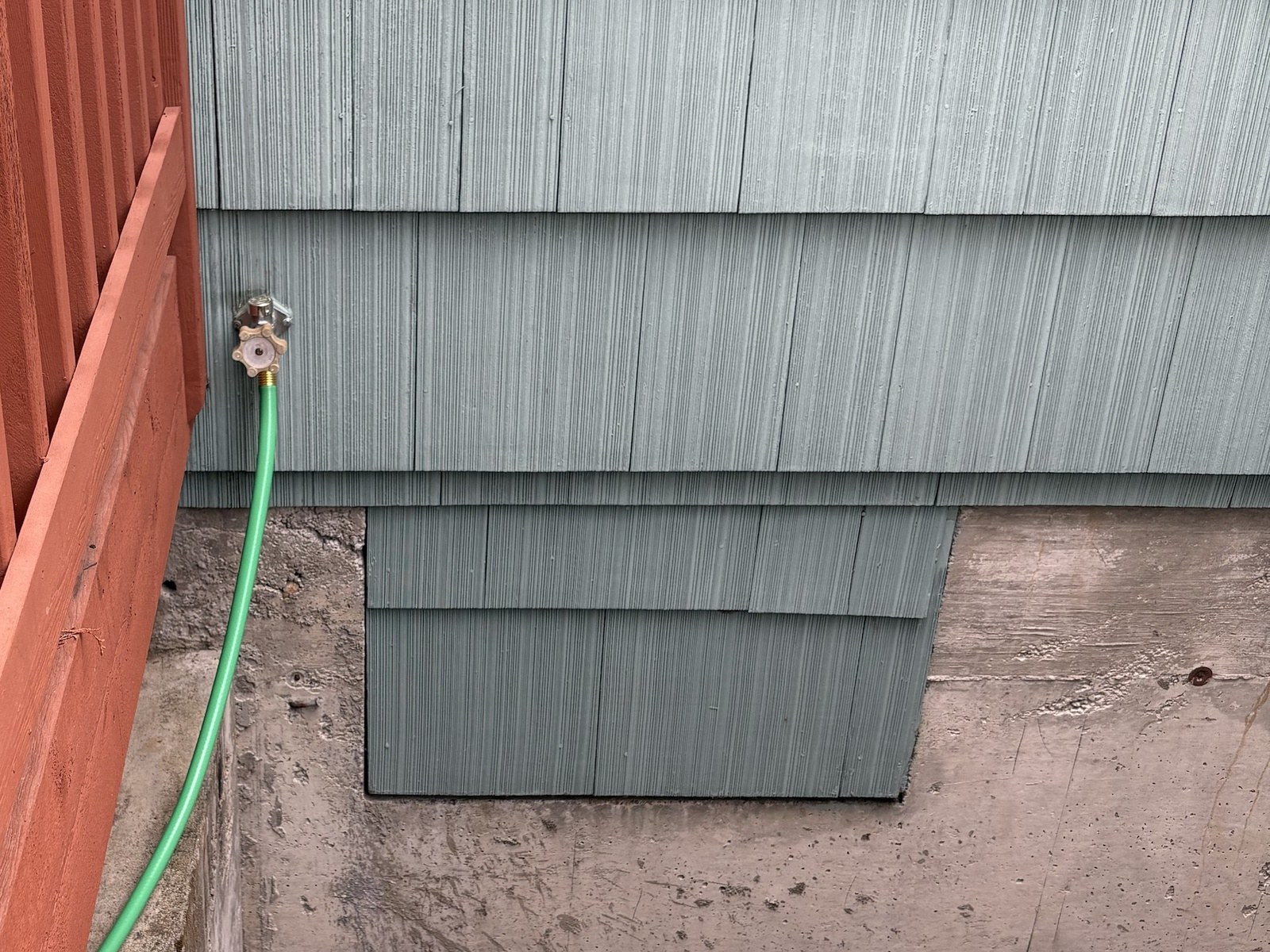 Outdoor faucet with a green garden hose attached, on blue-gray siding above a concrete basement stairwell beside a red deck railing