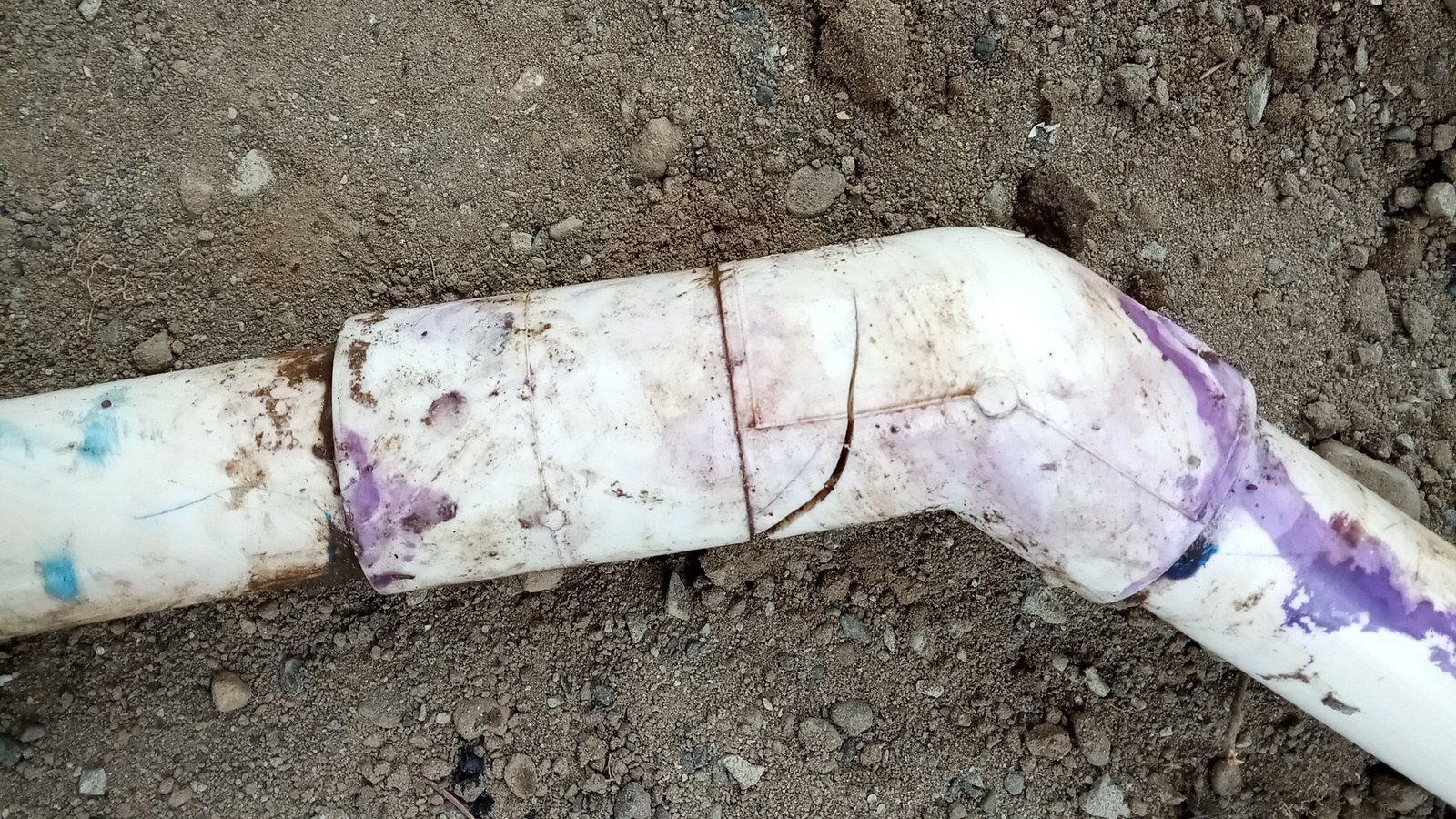White PVC pipe with a purple-primed elbow and coupling lying in brown soil, a thin crack running around the coupling