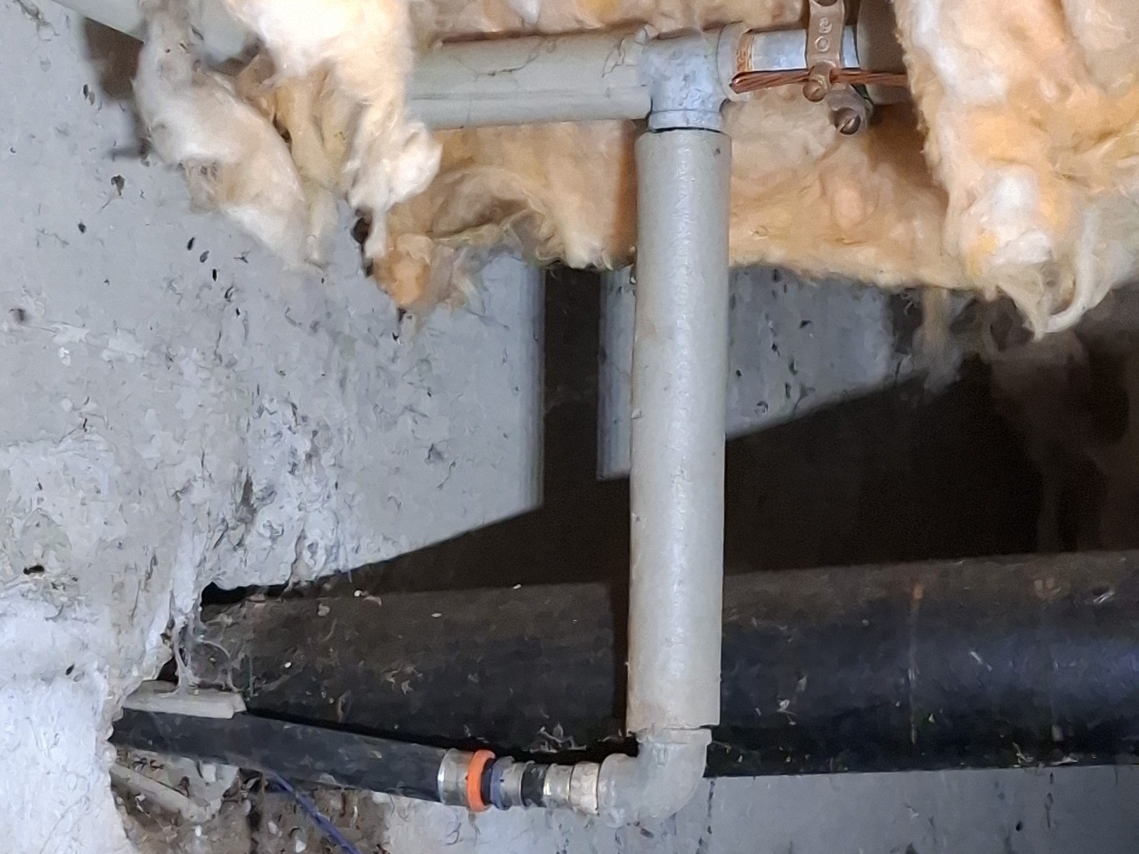 Crawlspace with a dark water service pipe entering through the concrete foundation and turning up into a galvanized riser and tee beneath fiberglass insulation