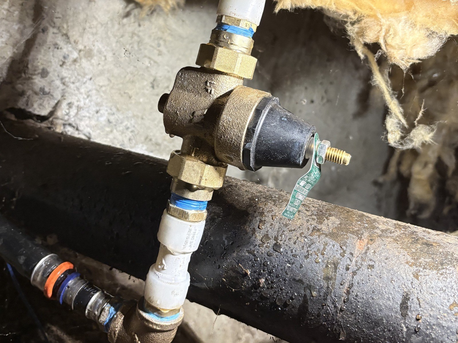 Brass pressure reducing valve with a black bell and a green tag installed upright on unions between white PEX pipes, above a large black pipe in a crawlspace