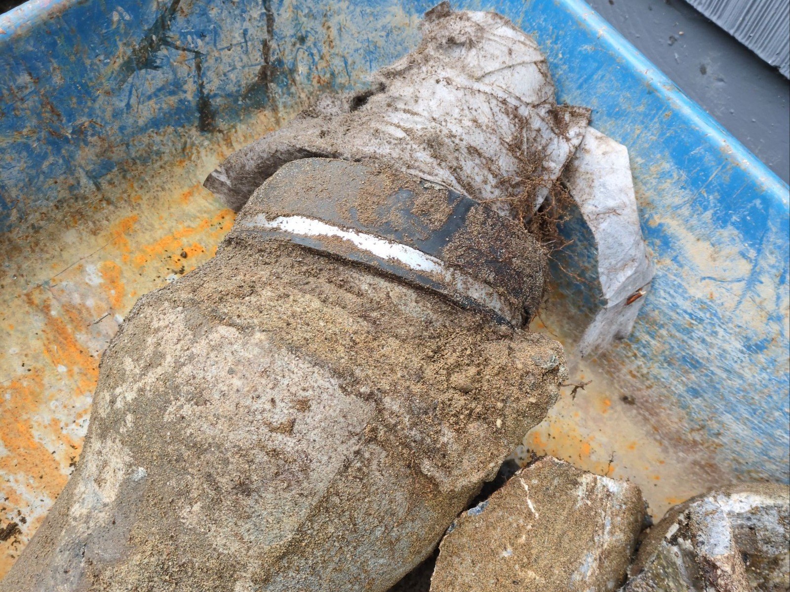 Broken concrete sewer pipe in a blue wheelbarrow, a cracked white plastic pipe jammed into its bell