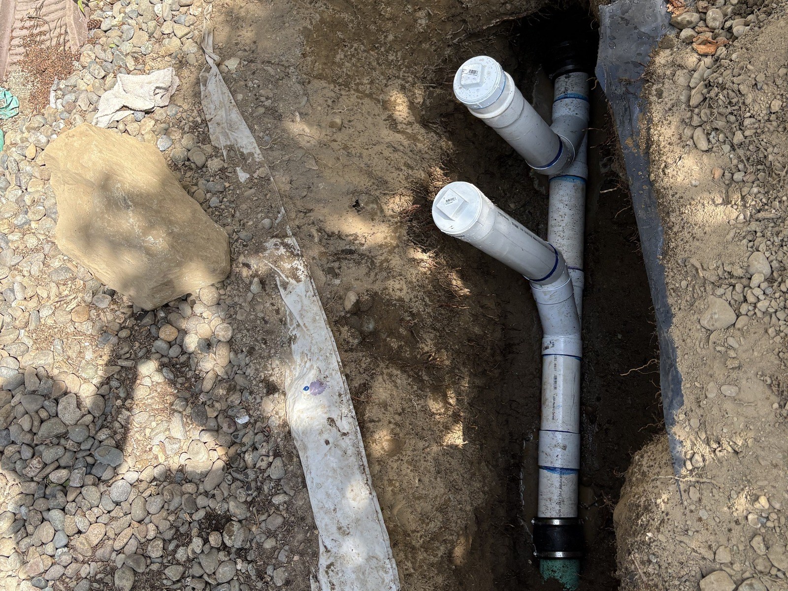 Open trench holding new white PVC sewer pipe, a wye and two capped cleanout risers, joined at the bottom to older green pipe with a black banded coupling