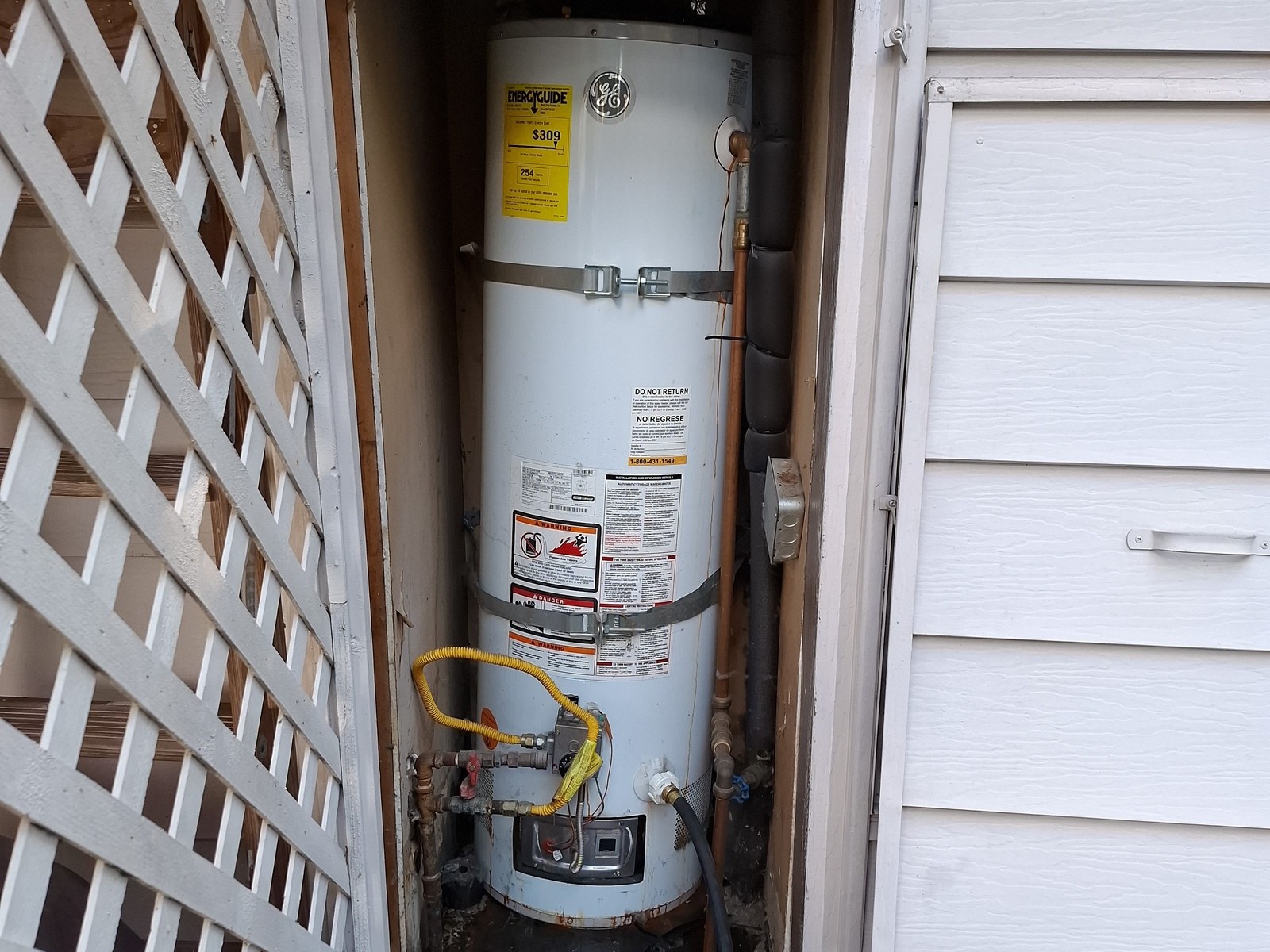 White gas water heater with a yellow gas connector and two metal straps, standing in a narrow closet behind an open lattice door in white siding
