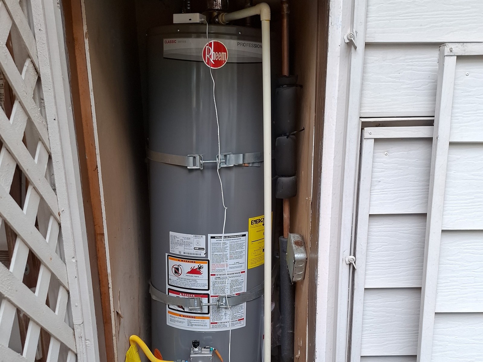 Gray Rheem gas water heater in the same narrow siding closet, with a white relief pipe running down its side and a yellow gas line at the burner