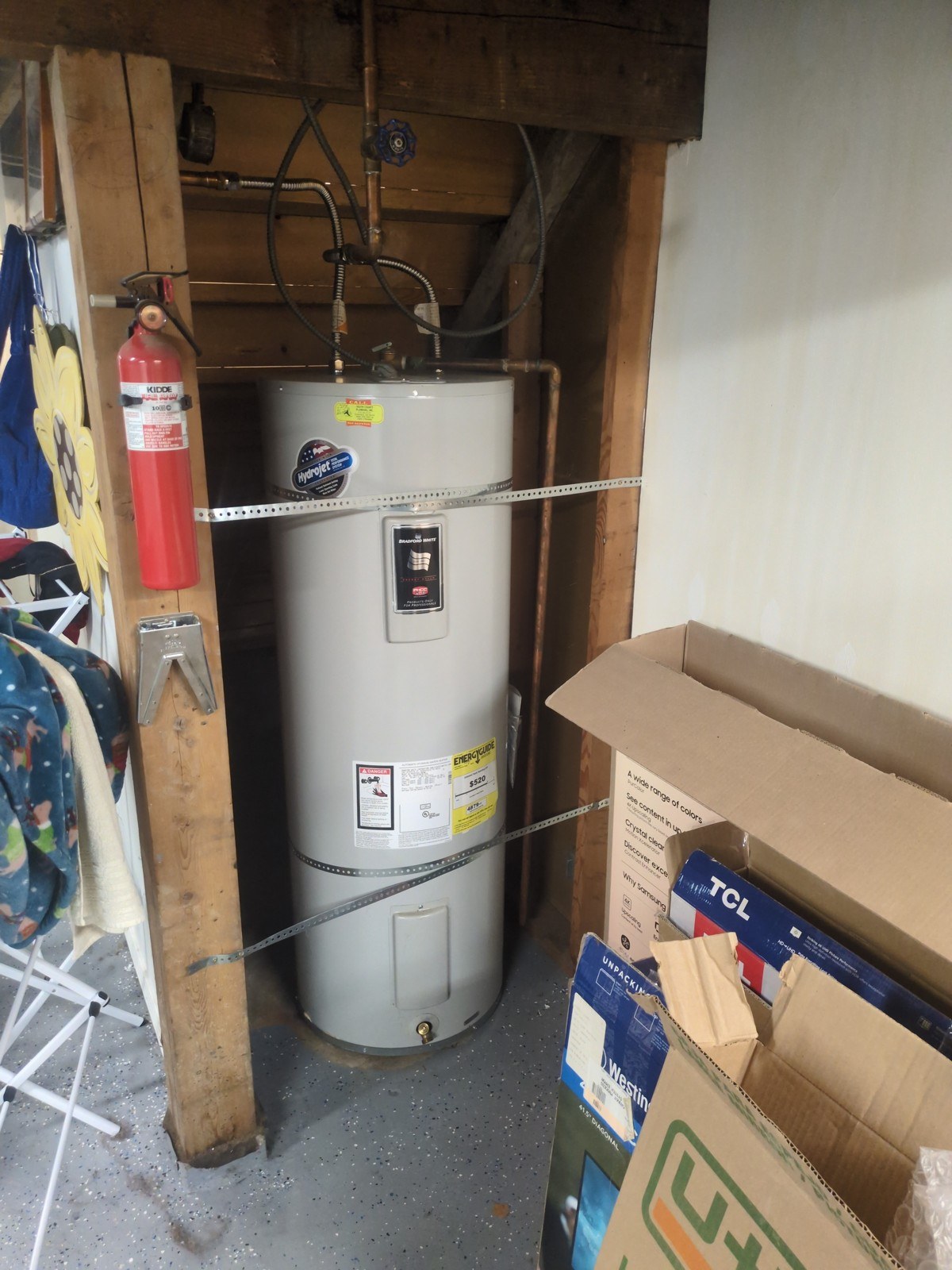 Old grey electric water heater in a Seattle garage between a wooden post with a fire extinguisher and stacked cardboard boxes, with perforated metal straps around its jacket