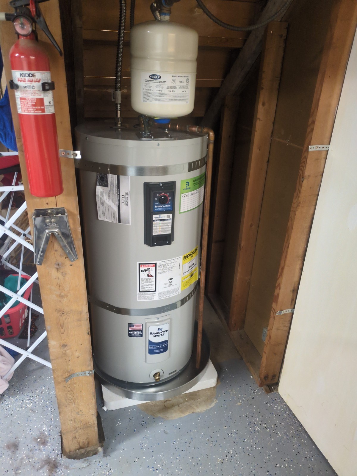 New Bradford White electric water heater in a Seattle garage with a white expansion tank mounted above it, two new seismic straps, a copper relief line running down the side and a dated service sticker on the jacket