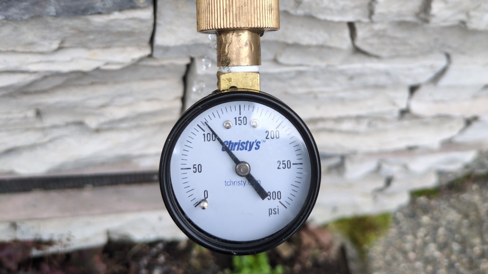 Brass-fitted test gauge with its needle past the 100 mark, threaded onto a green-handled hose bib against stacked stone siding