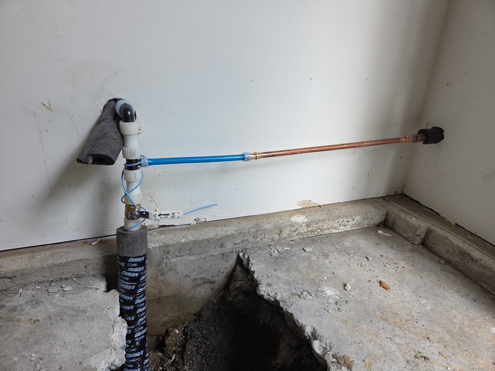 New black PEX water pipe rising out of a hole cut in a garage floor, wrapped in tape and foam, with a new ball valve and a blue line running along the wall