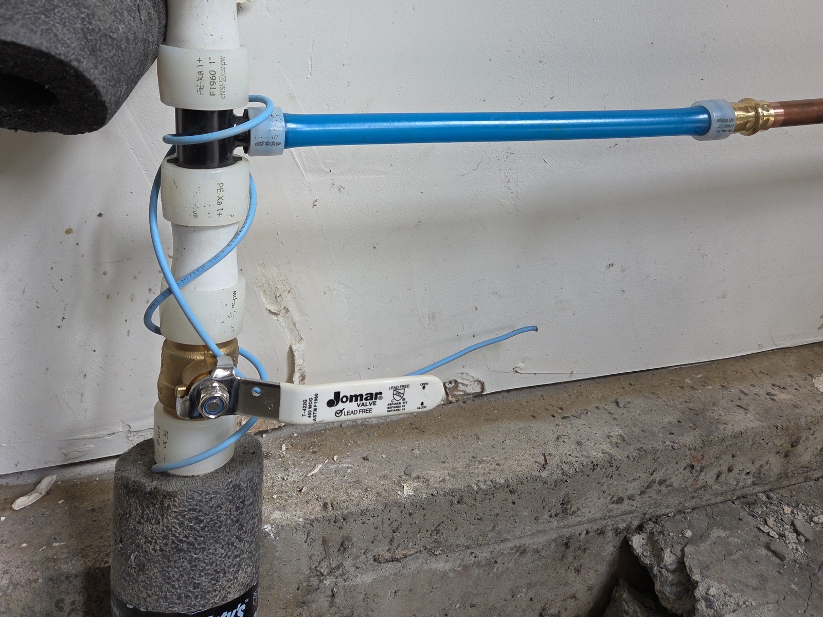 Close view of a new white-handled quarter-turn ball valve on PEX pipe against a garage wall, with blue PEX branching off toward copper