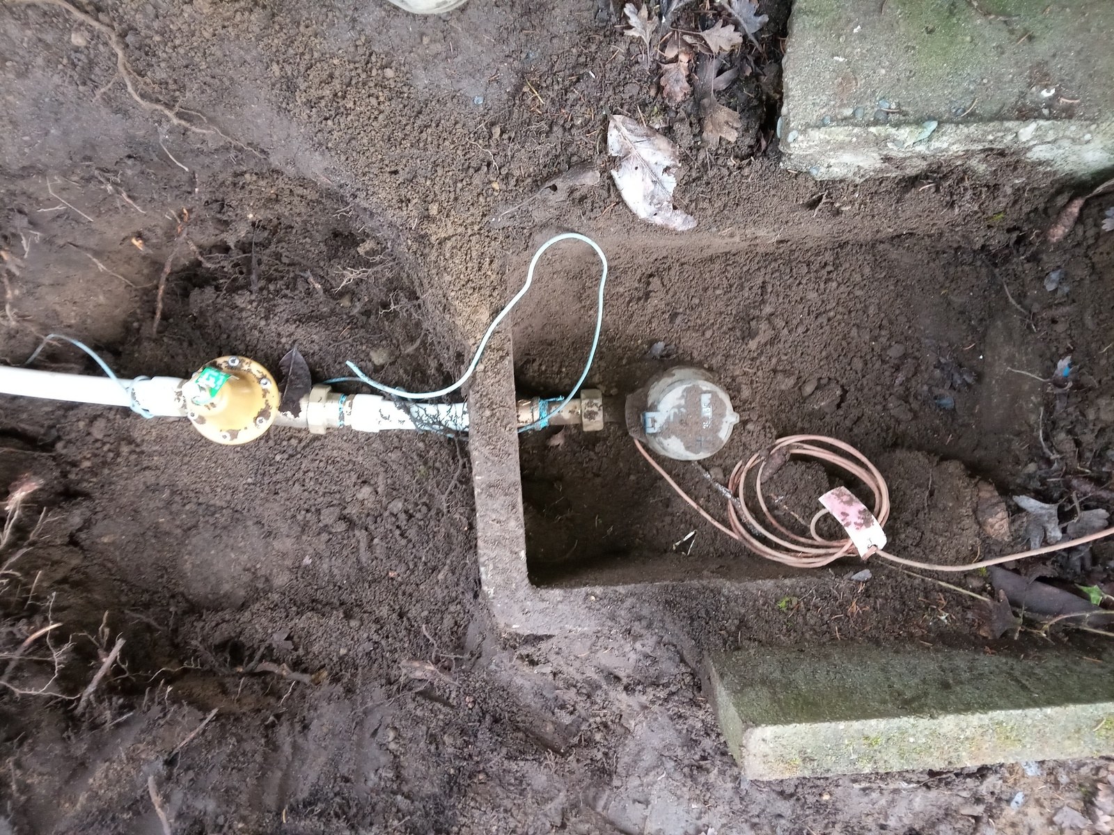 Excavated trench showing a new water service line running toward a concrete meter box with a coiled tracer wire