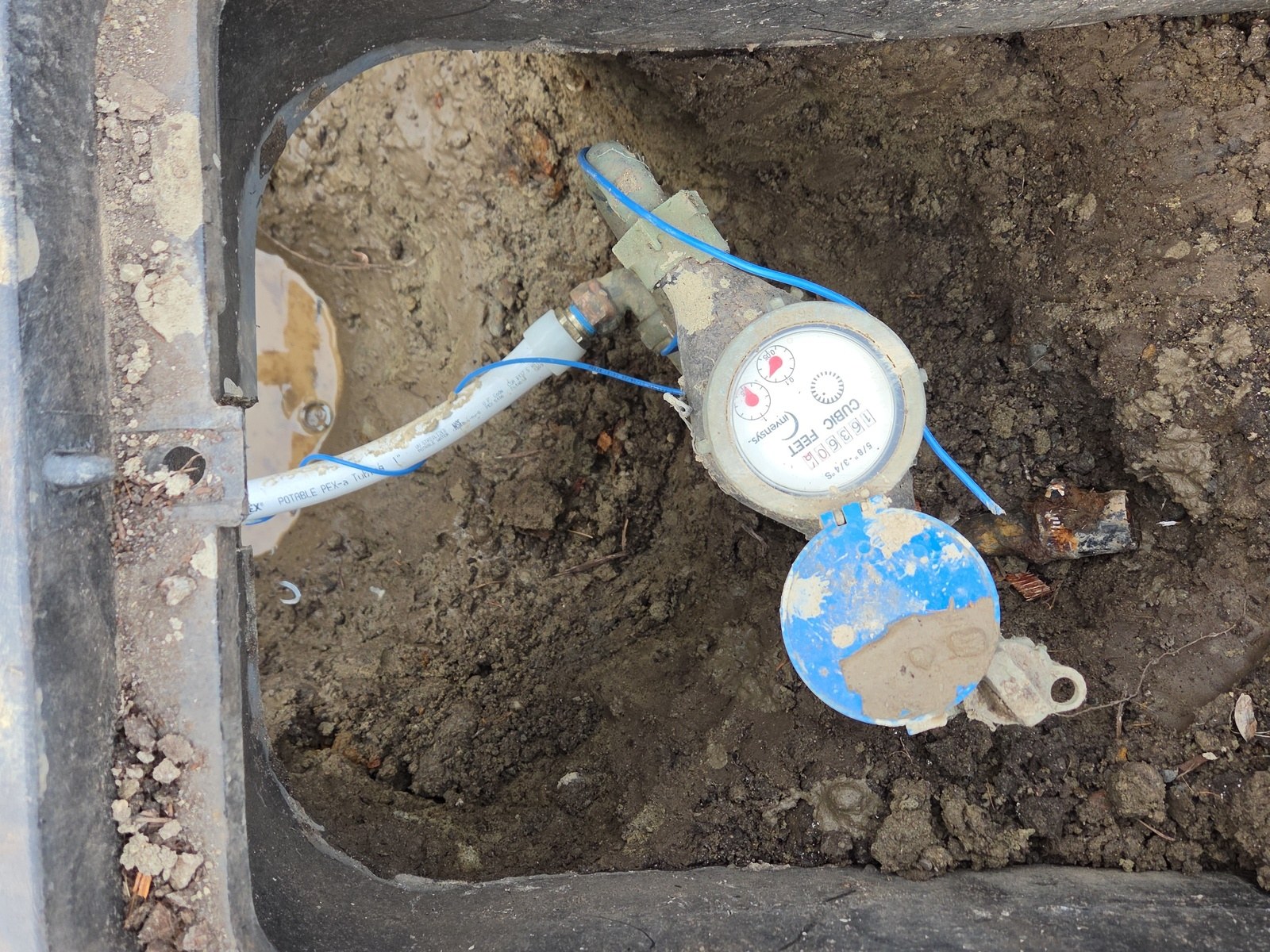 Water meter in an open meter box, with white PEX pipe connected at its outlet by a brass fitting and a thin blue wire looped beside it