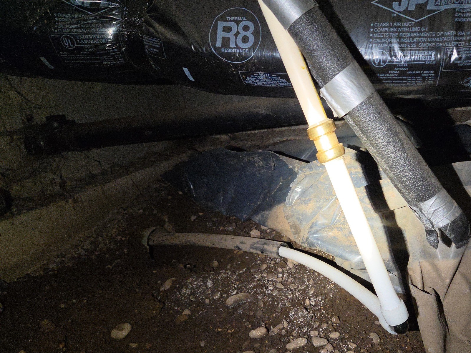 White PEX A pipe emerging from the end of an older pipe at a crawlspace footing, turning up through an elbow to a brass coupling beside a foam-insulated line under flexible ductwork