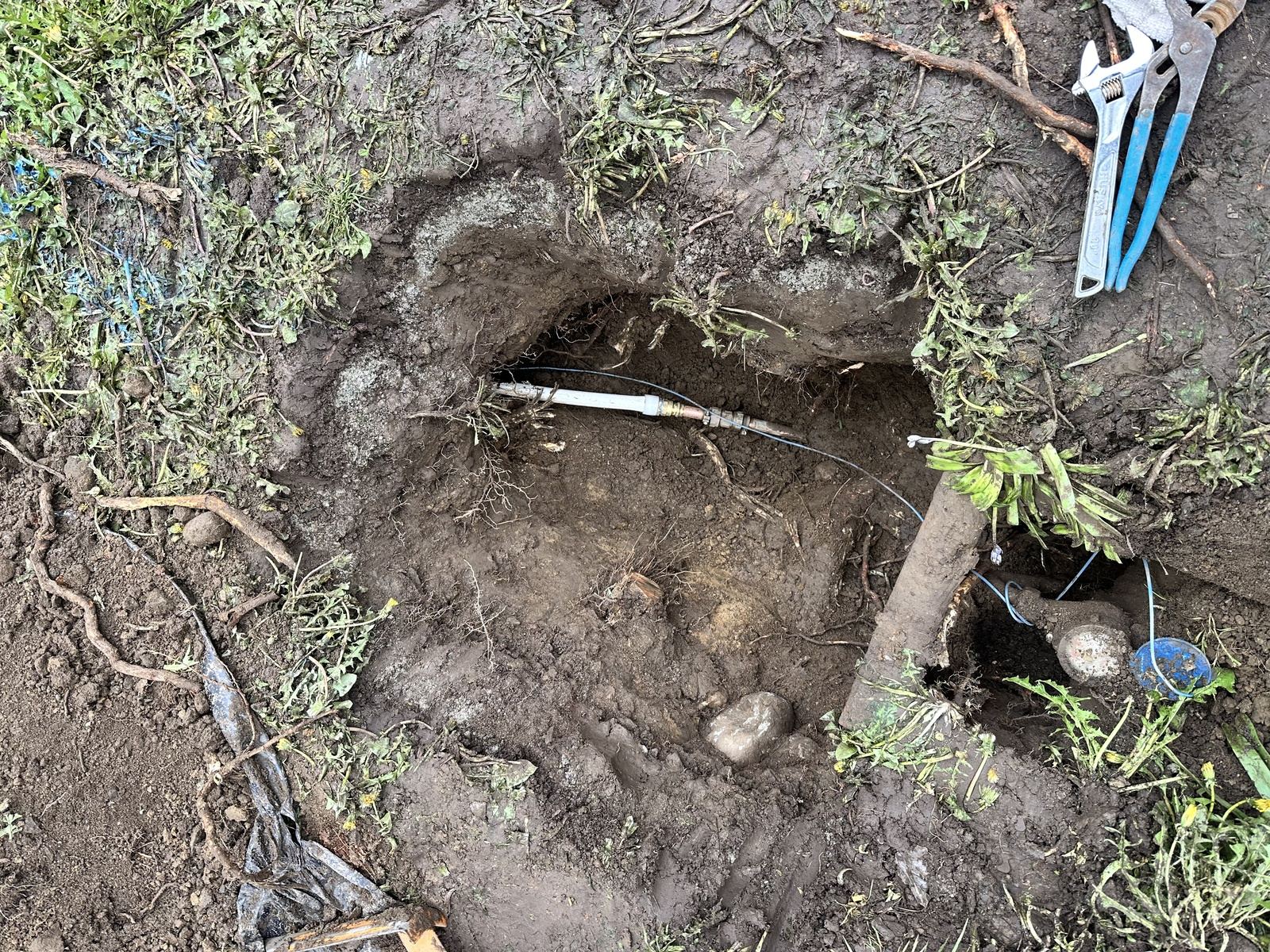 Hand-dug pit showing a new white water service line joined to copper with brass compression fittings, blue tracer wire alongside and the meter box at the edge