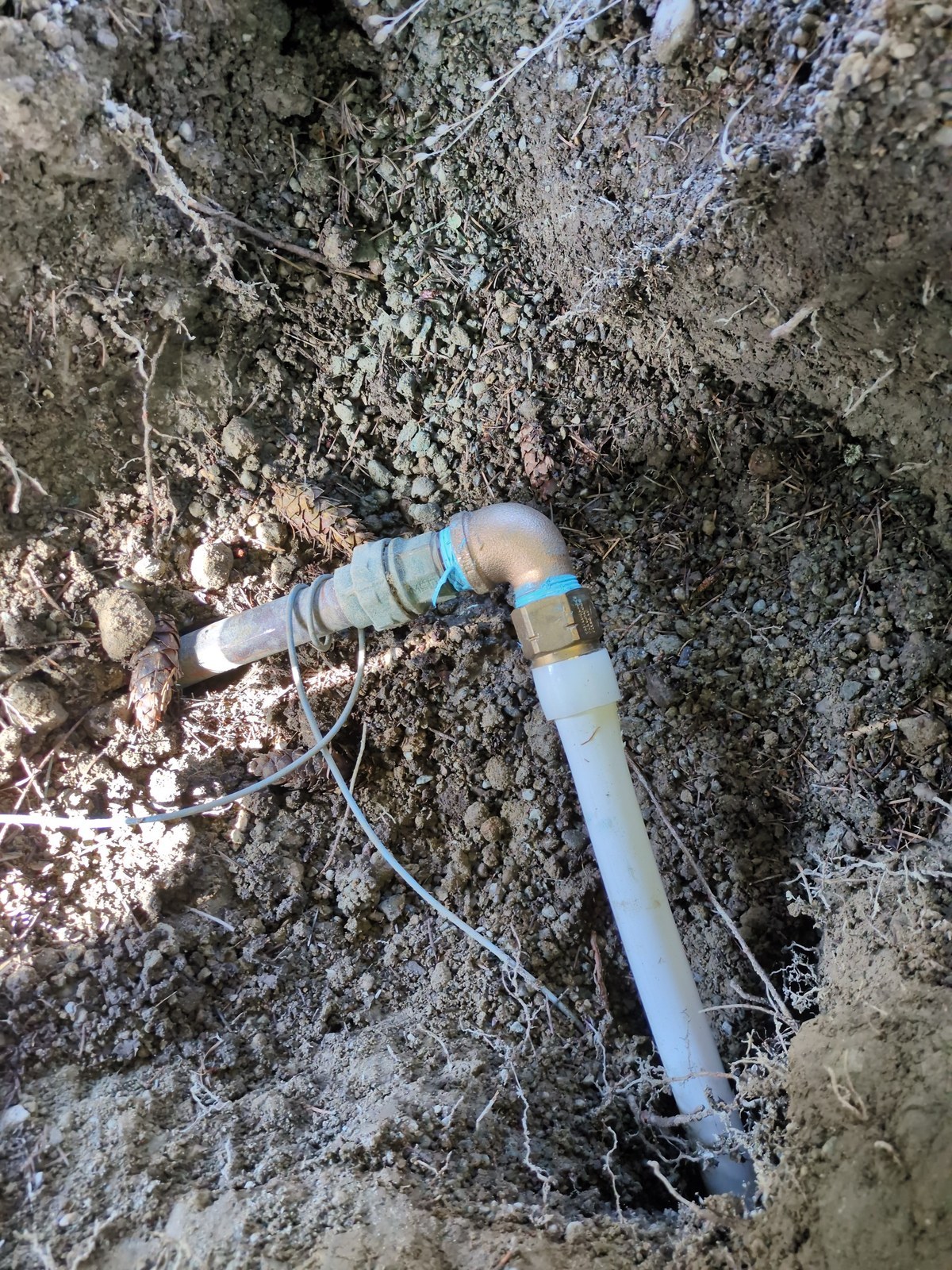 Close-up inside a dirt pit of an old galvanized water line meeting a brass elbow and compression fitting, with new white PEX A pipe and a blue tracer wire running away from it