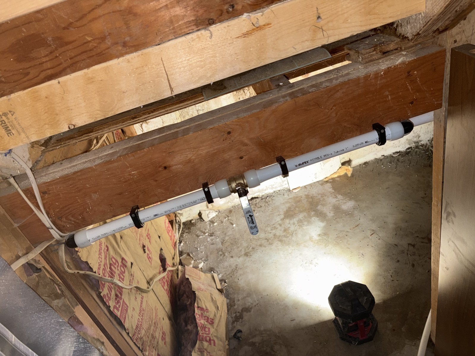 White one-inch PEX A tubing clipped to a wooden beam in a crawlspace, with a brass quarter-turn ball valve in the run and kraft-faced insulation behind it