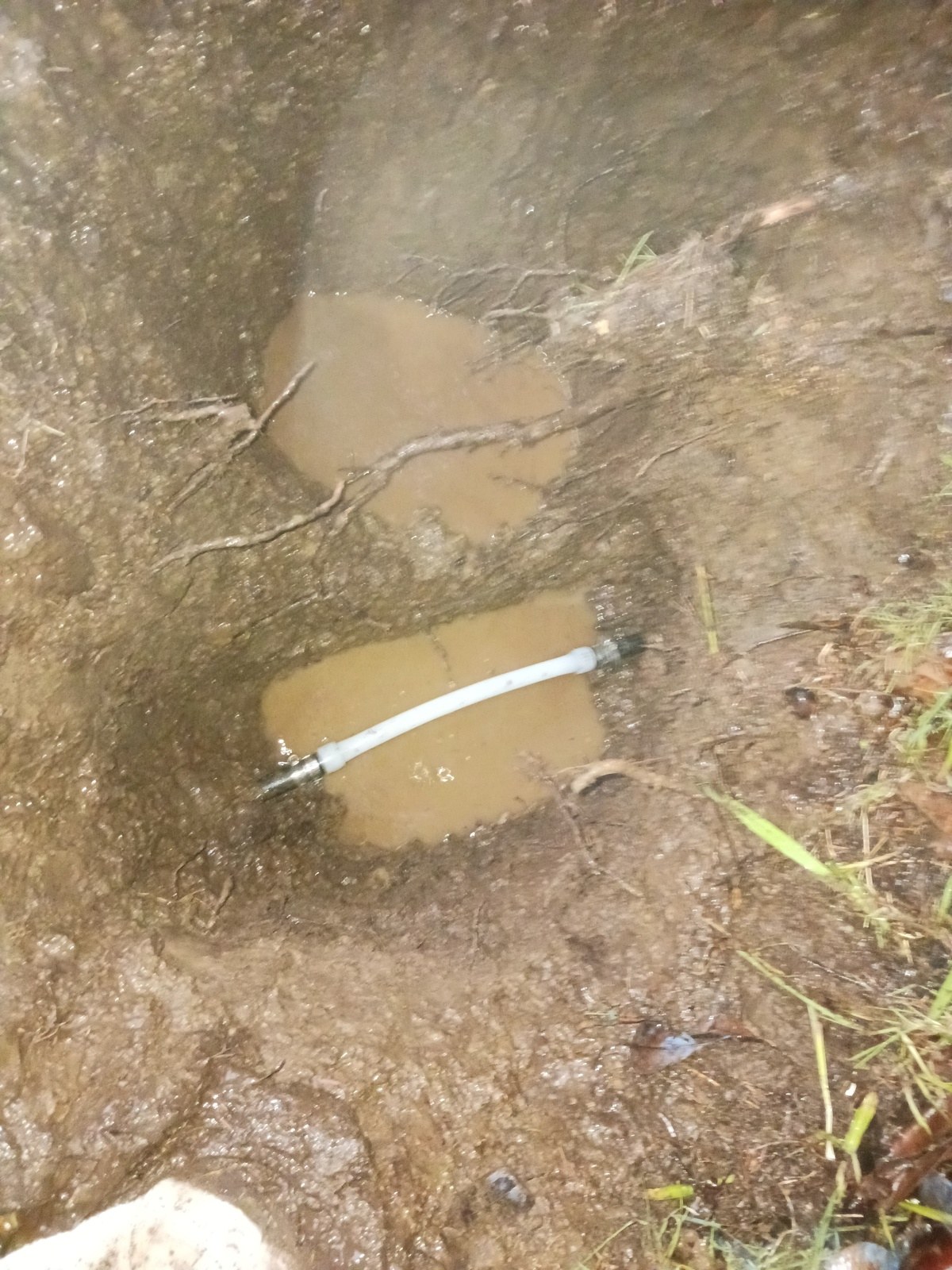 Open repair pit with a white spliced water line crossing muddy standing water, wrenches at the edge of the excavation